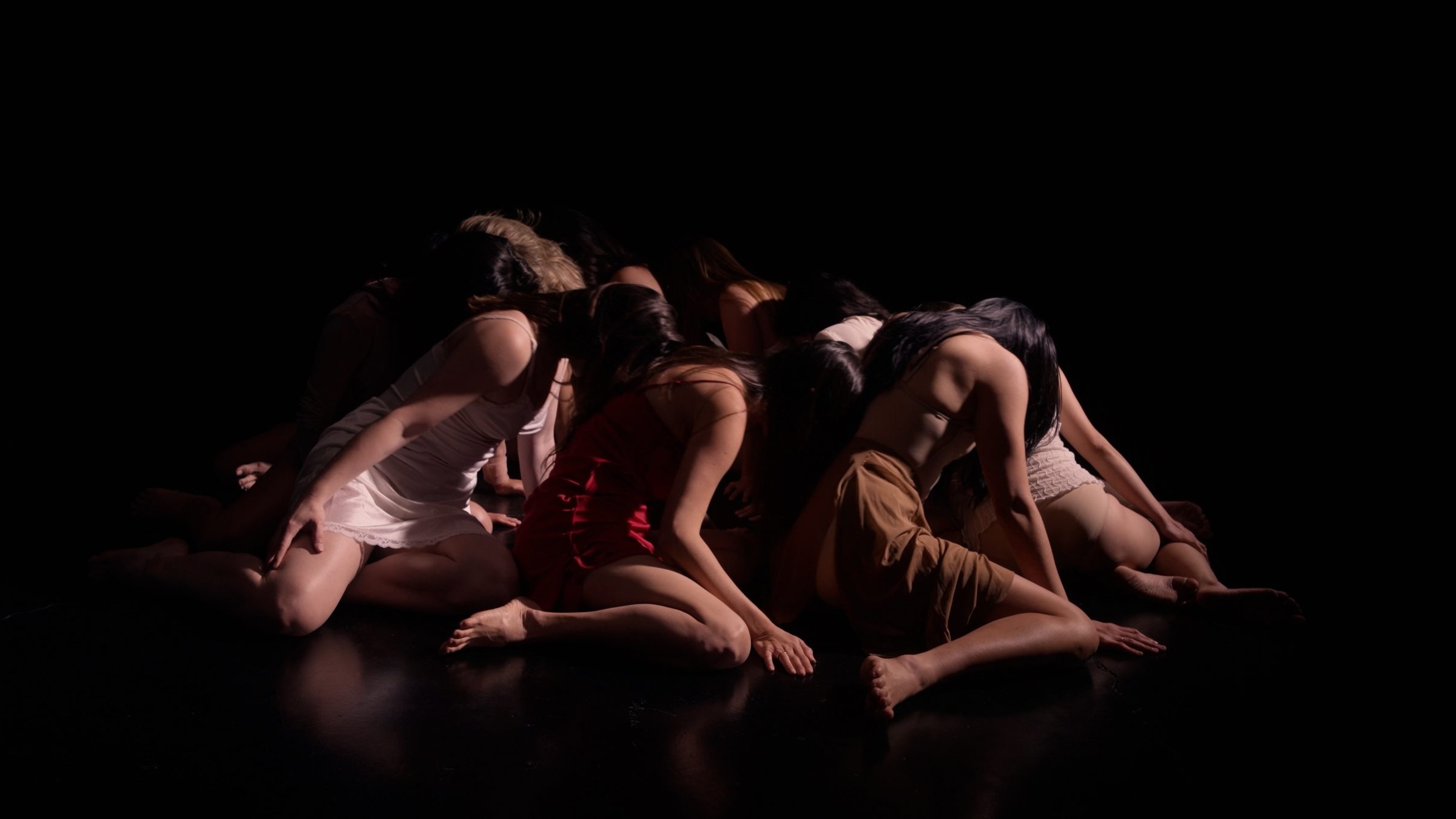 Film still: a group of dancers huddled together on a dark stage floor